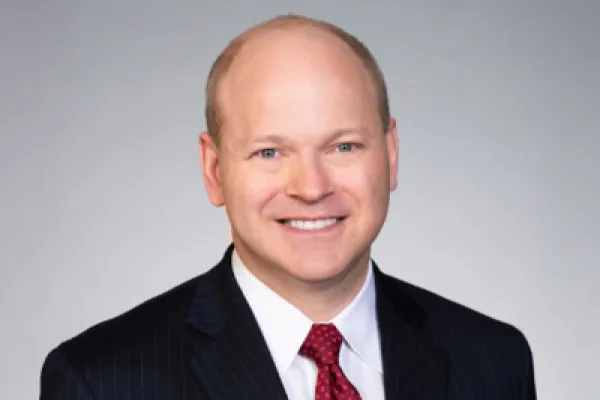 A man in a dark pinstripe suit with a red tie against a plain gray background.