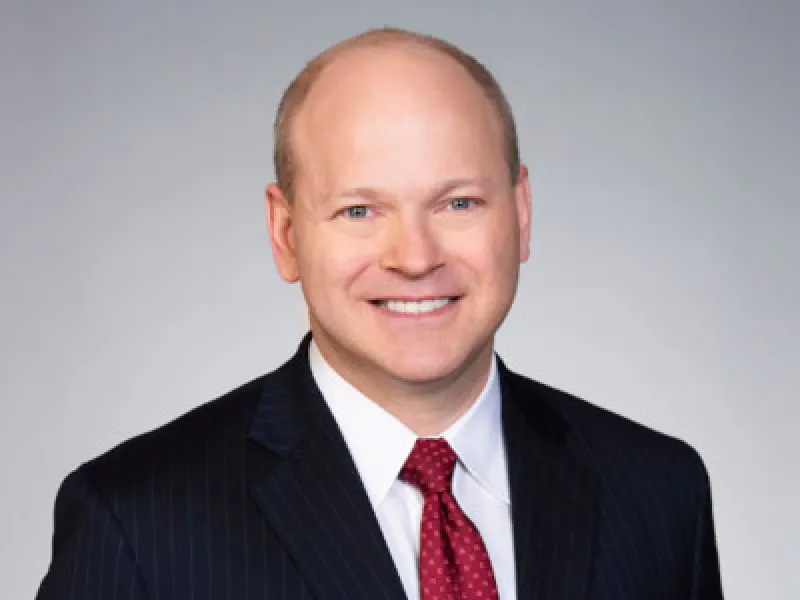 A man in a dark pinstripe suit with a red tie against a plain gray background.