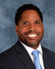 A man with dark hair in a suit, tie, and white shirt against a blue background.